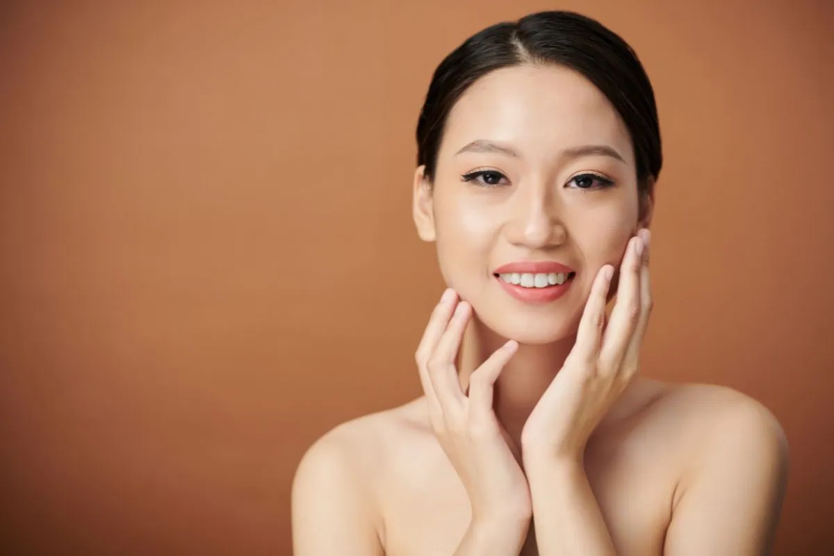 Smiling Asian woman with radiant, clear skin touching her face.