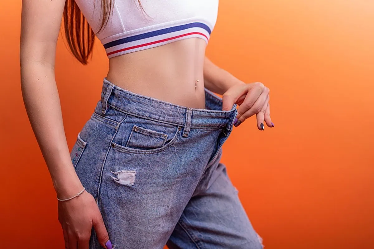 Slim woman with a toned abdomen shows off her weight loss in oversized jeans.