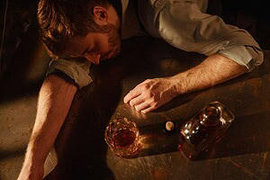 Man passed out on a metal table next to a glass and bottle of liquor.
