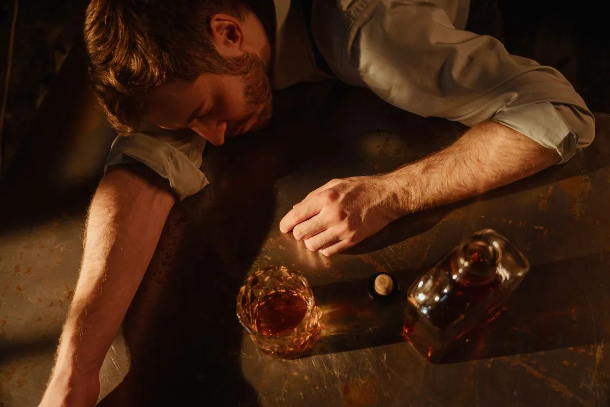 Man passed out on a metal table next to a glass and bottle of liquor.