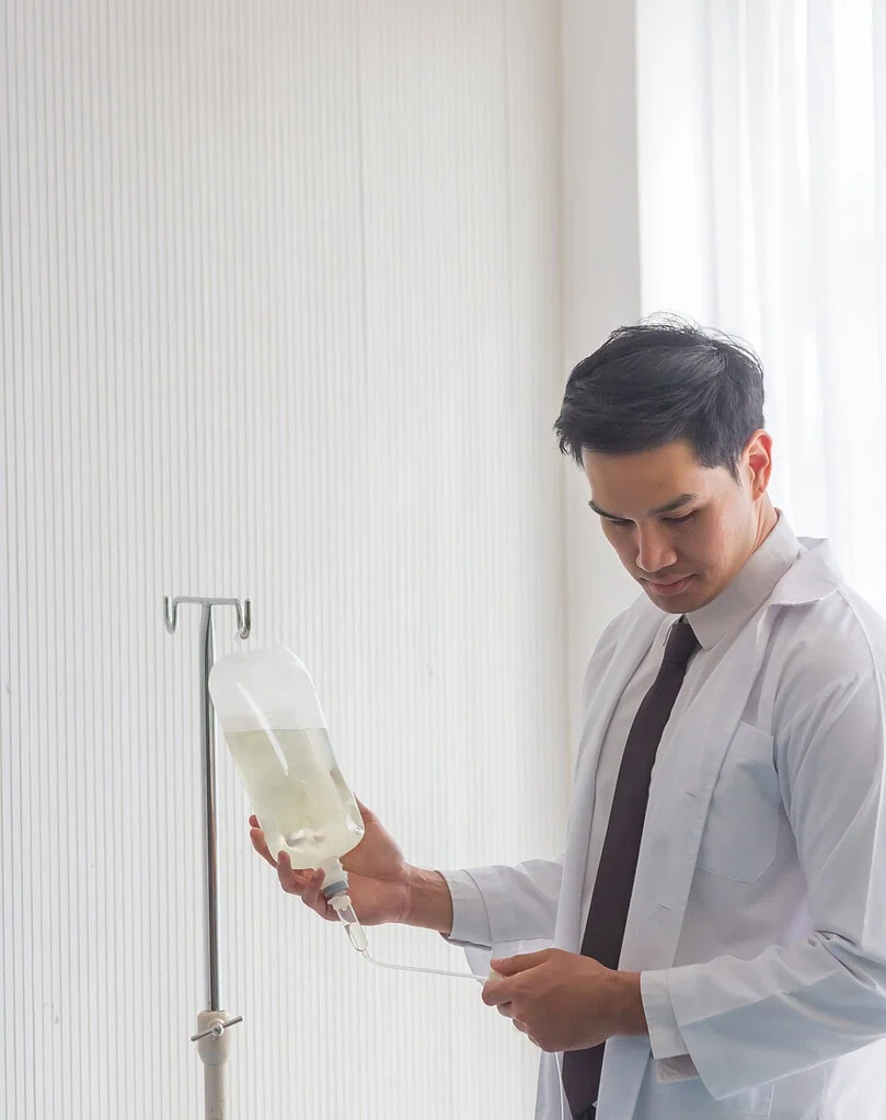 Male doctor in white lab coat checking an IV fluid bag on a stand.