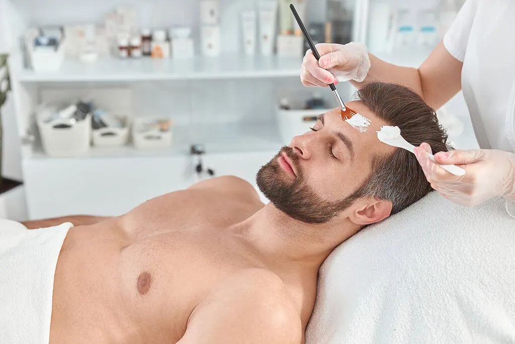 Man receiving a white facial mask applied with a brush by a therapist.