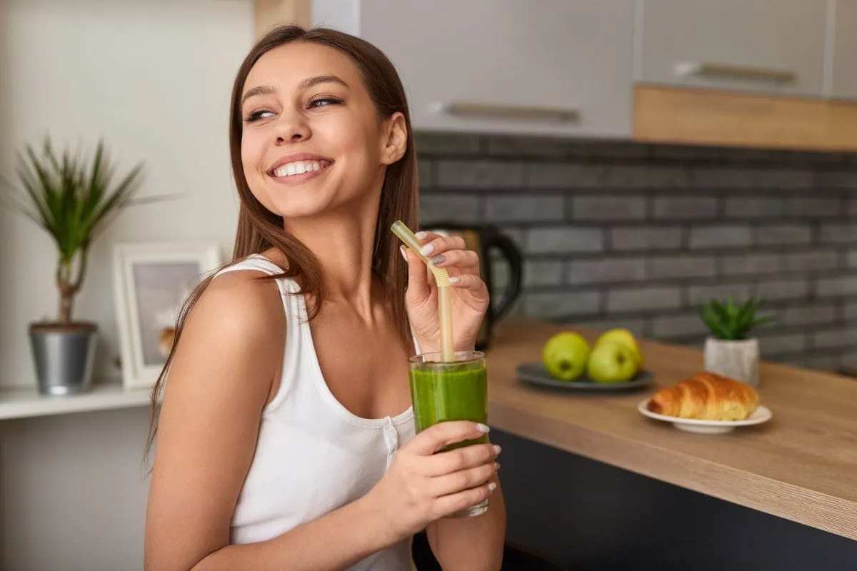 Smiling woman holding a glass of green smoothie in a kitchen.
