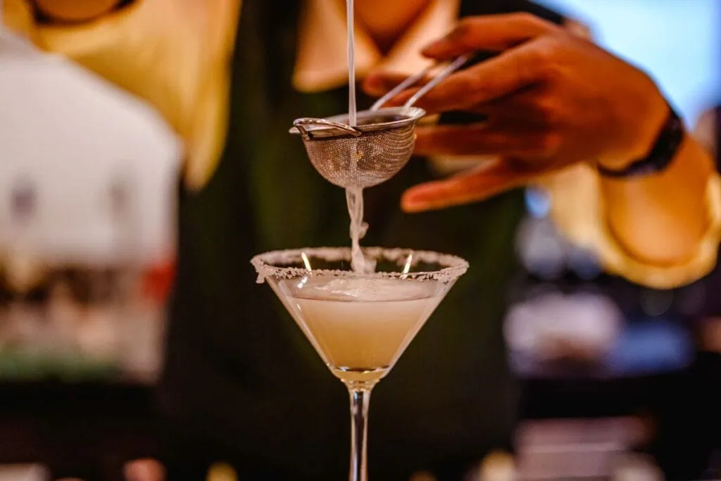 Bartender pouring cocktail through a strainer into a rimmed glass.