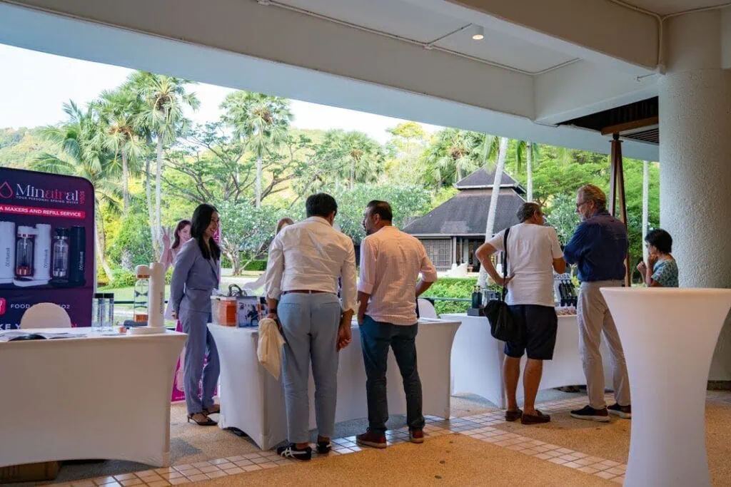 A group of people gathered around a table at an outdoor event, with lush greenery and palm trees in the background, under a covered patio at Pullman Phuket Karon Beach Resort.
