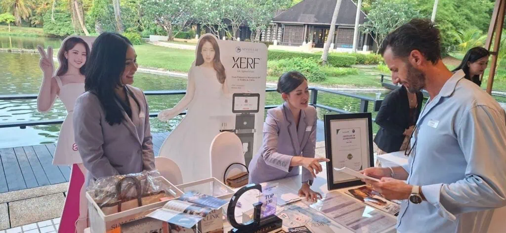 A woman in a gray blazer and a man in a light blue shirt exchanging information at an outdoor event booth with promotional materials, near a water feature and greenery.