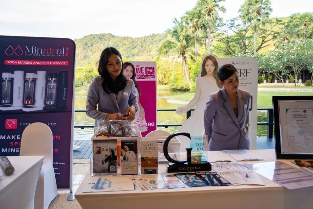 A woman and a girl in gray uniforms standing at a promotional booth outdoors, with banners and product displays, during the Siam Clinic event at Pullman Phuket Karon Beach Resort.