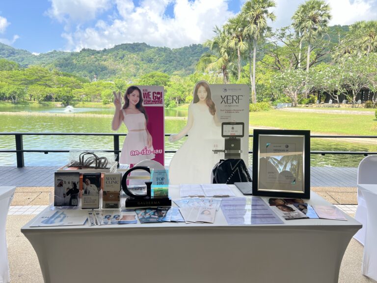 A table displaying promotional materials and products for Siam Clinic at an outdoor event with a lake, trees, and mountains in the background.