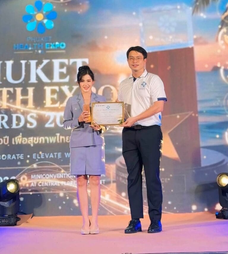 Two individuals on stage at Phuket Health Expo 2026, one woman holding a certificate and a man standing beside her, both smiling with a large event banner in the background.