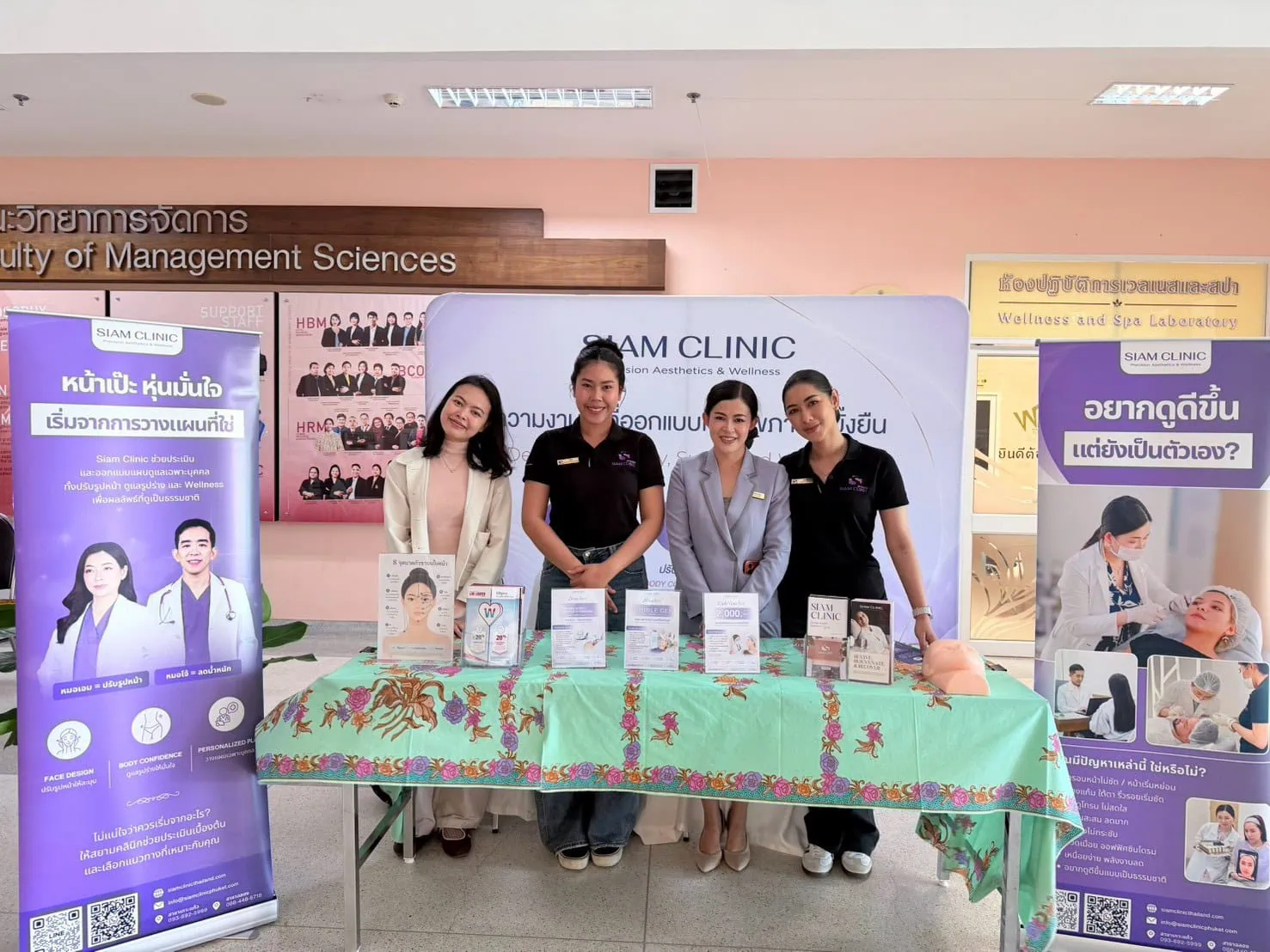 Four women stand behind a table with informational materials at Siam Clinic's Wellness Day event in a university corridor.