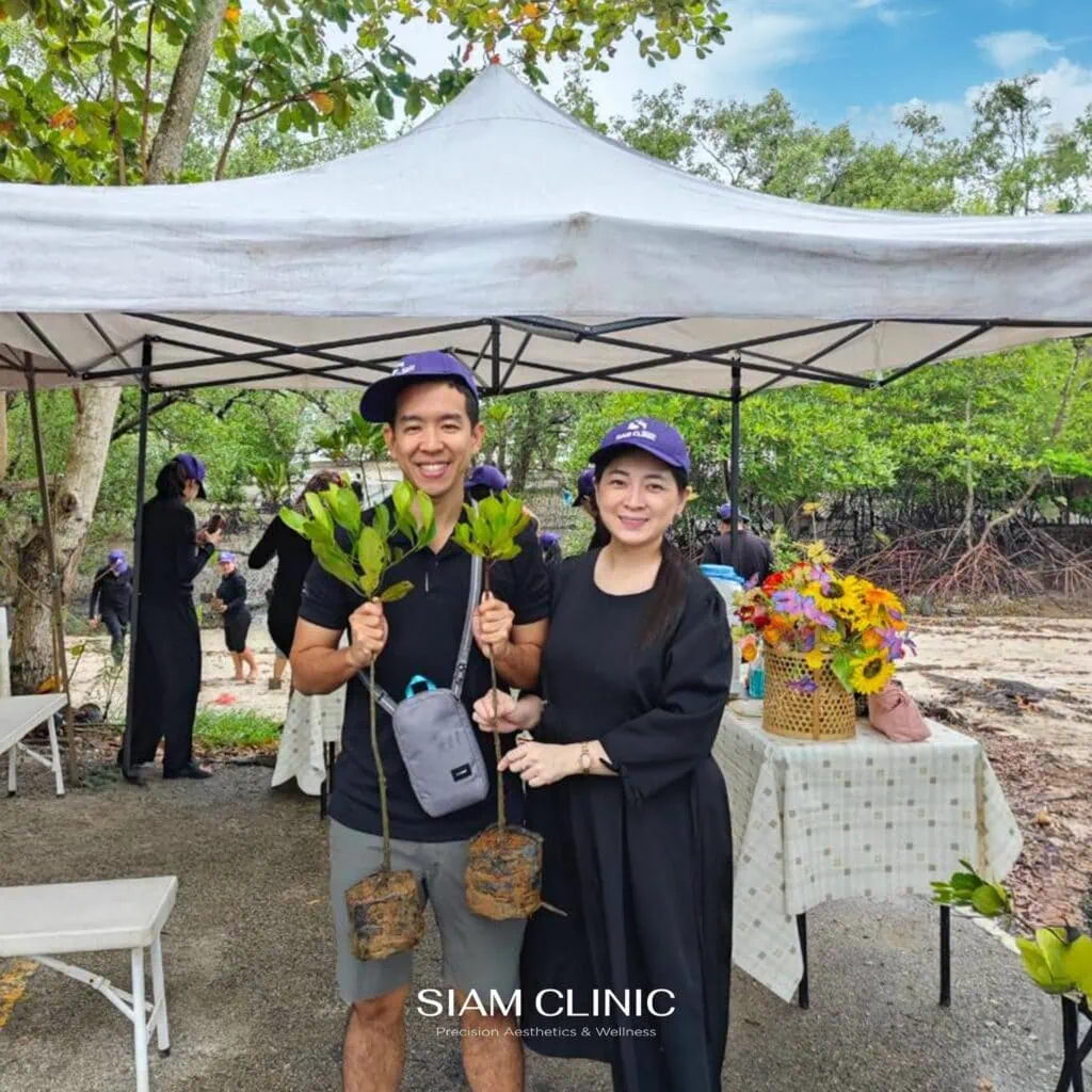 Two smiling women in black shirts and purple caps holding young mangrove saplings at a tree planting event under a white canopy with a table of flowers in the background.