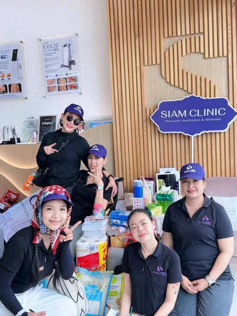 Five staff members in black uniforms and blue caps pose behind a table with health products at Siam Clinic, with a wooden wall and clinic sign in the background.