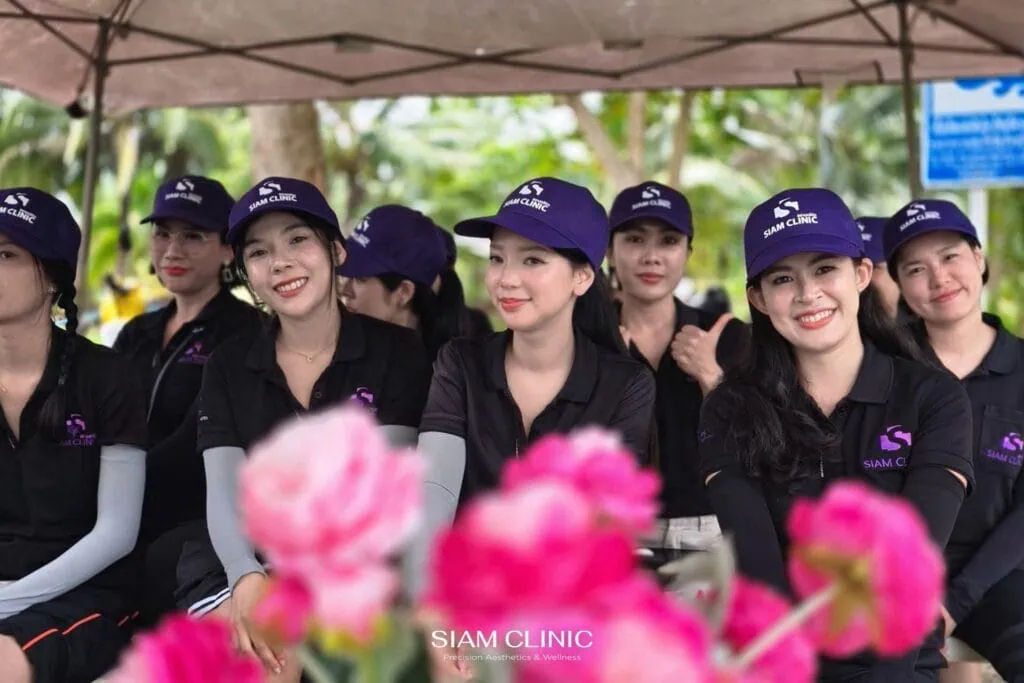 Group of women wearing black shirts and purple caps with Siam Clinic logo, sitting outdoors under a tent, smiling at the camera with pink flowers in foreground.
