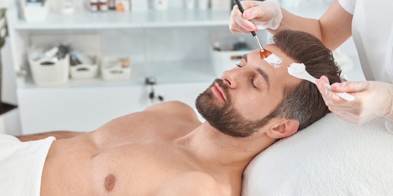 Man receiving a white facial mask applied with a brush by a therapist.